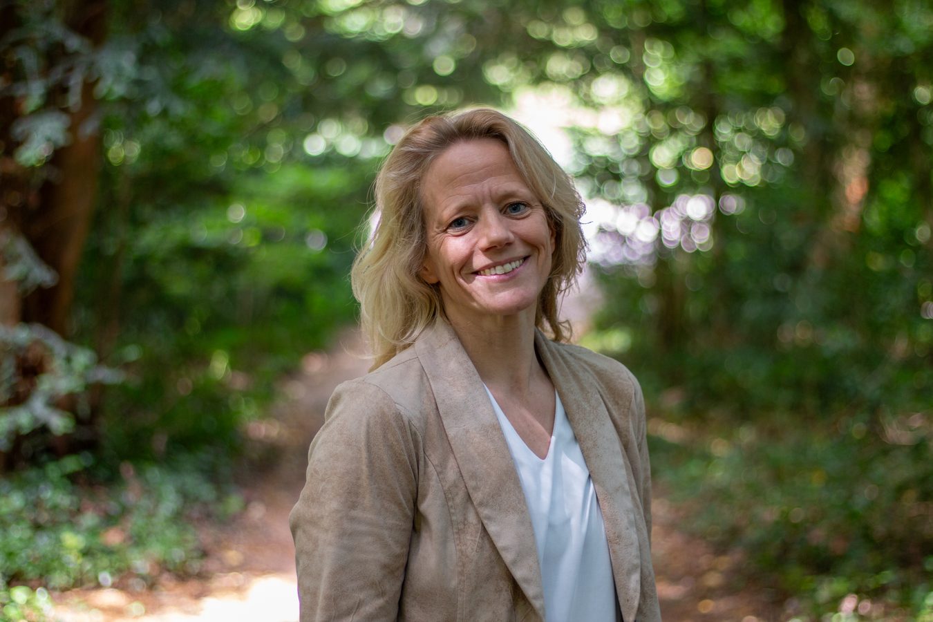 Leende kvinna på en skogsstig omgiven av grönska en solig dag. Kvinnan är blond och har en beige kavaj över vit v-ringad t-shirt.