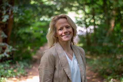 Leende kvinna på en skogsstig omgiven av grönska en solig dag. Kvinnan är blond och har en beige kavaj över vit v-ringad t-shirt.