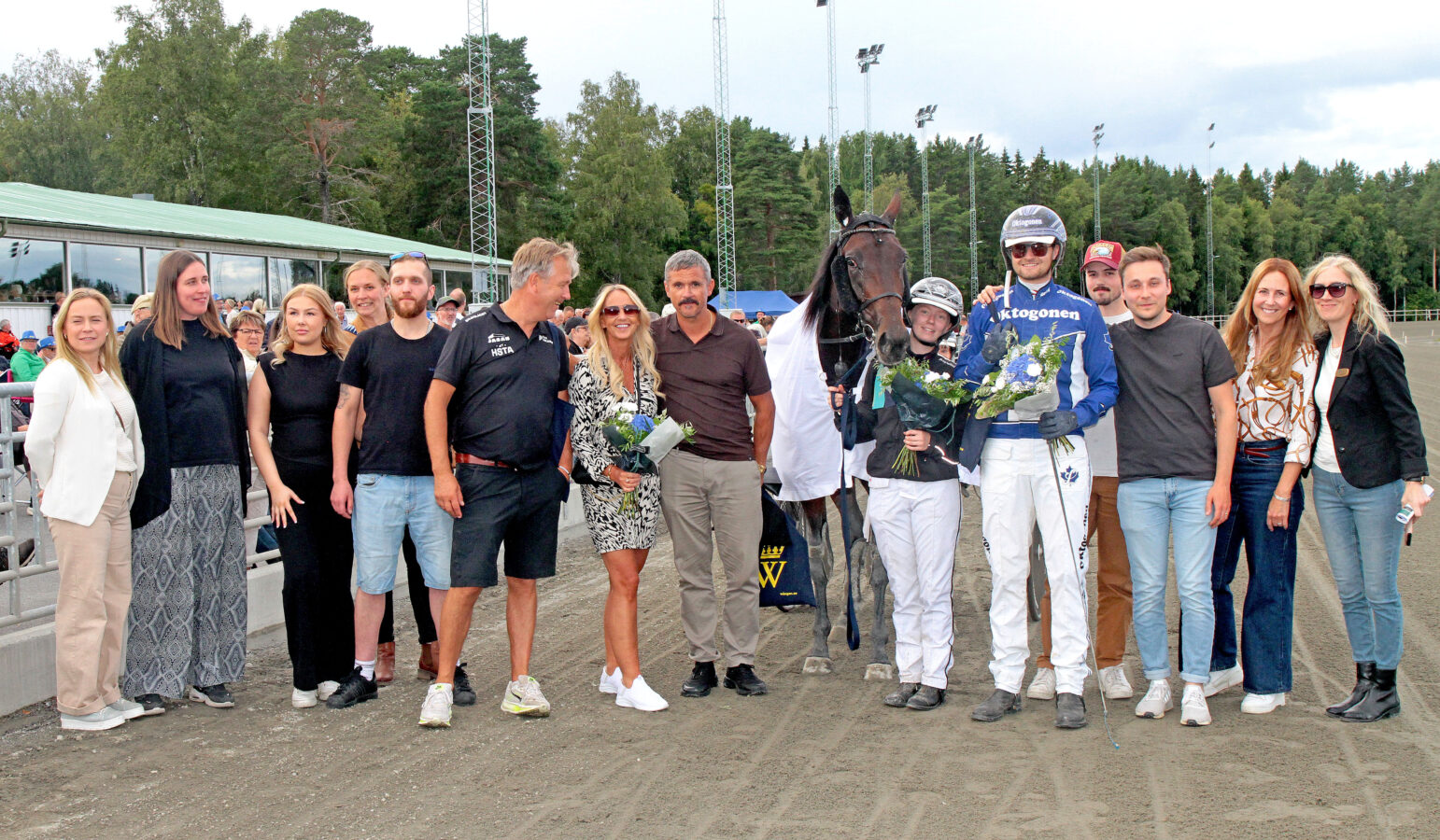Gruppfoto på travbana med häst och personerna runt hästen och loppets sponsorer. Kusken, hästägaren och skötaren håller blombuketter.