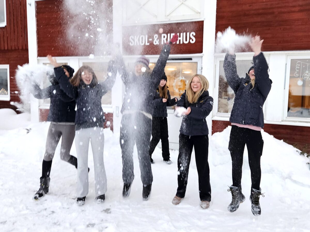 Elever utanför en röd byggnad med skylten Skol- och ridhus på kastar snö upp i luften och ser glada ut