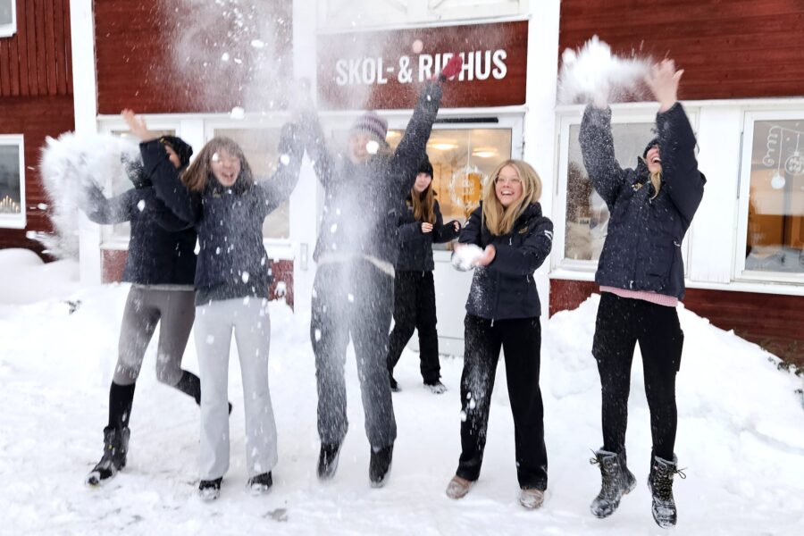 Elever utanför en röd byggnad med skylten Skol- och ridhus på kastar snö upp i luften och ser glada ut