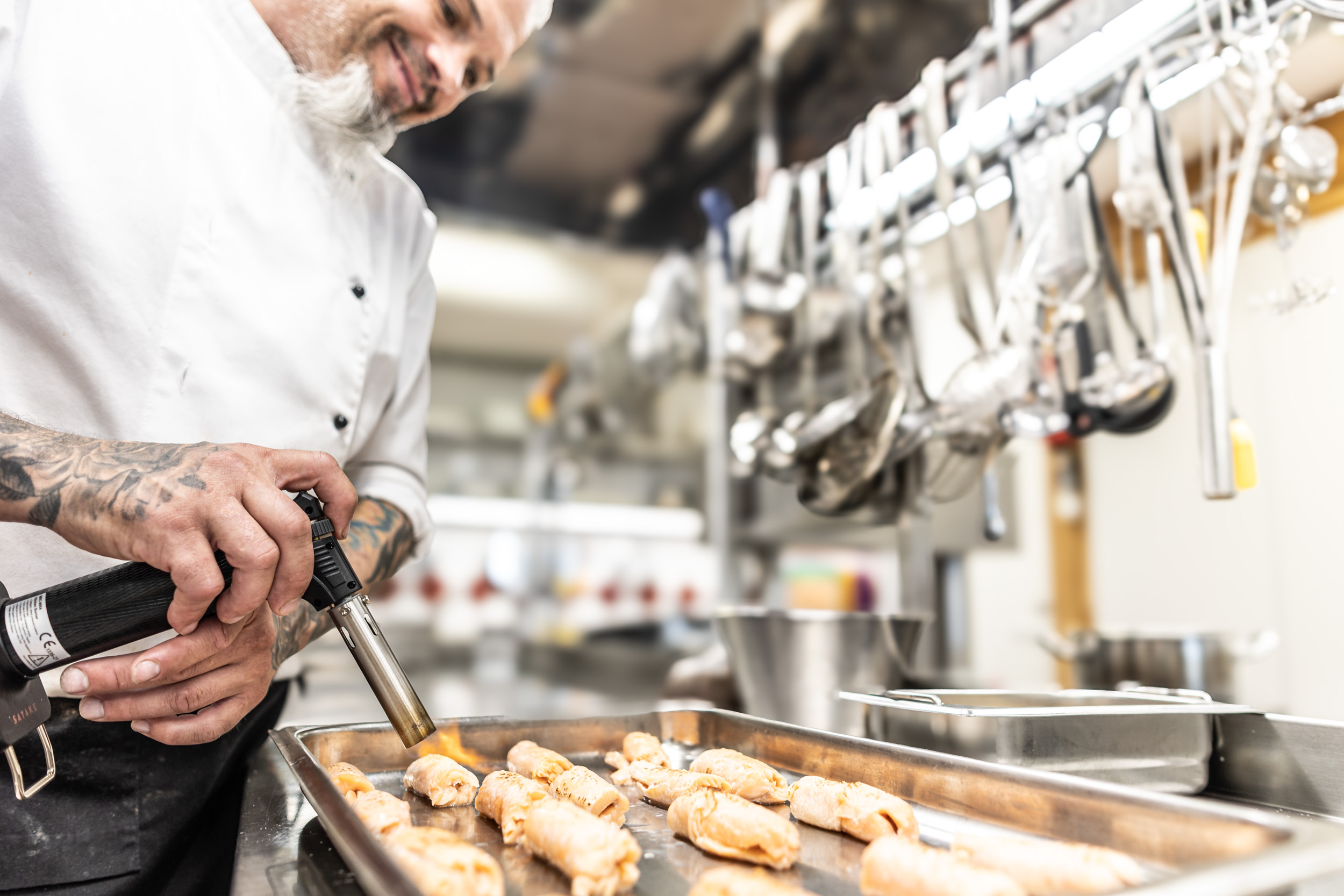 I ett livligt restaurangkök använder kocken skickligt en blåslampa på bakverk, omgiven av hängande redskap.