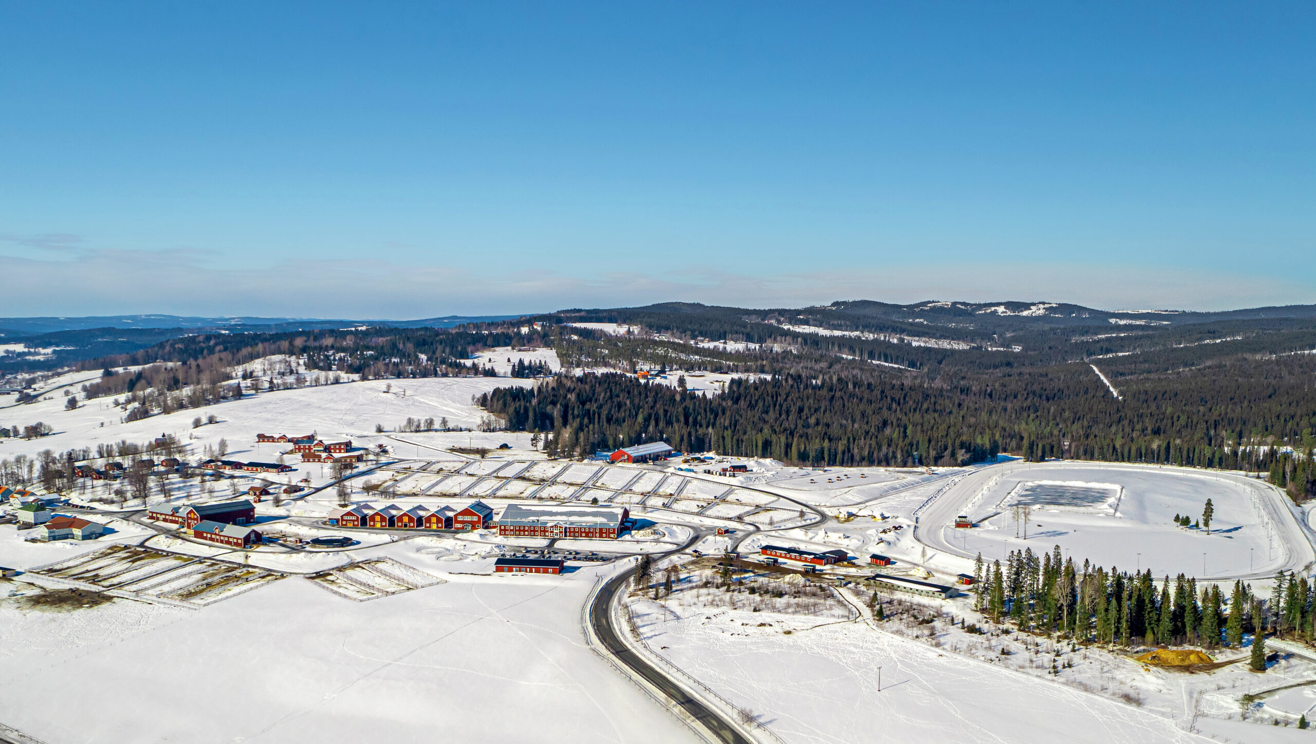 En flygvy över det snöiga landskapet runt Wången med spridda byggnader, slingrande vägar och ett skogsområde under en klarblå himmel.