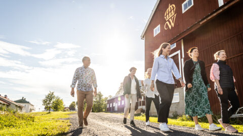 En grupp människor går på en grusgång bredvid en röd lada under en solig himmel.