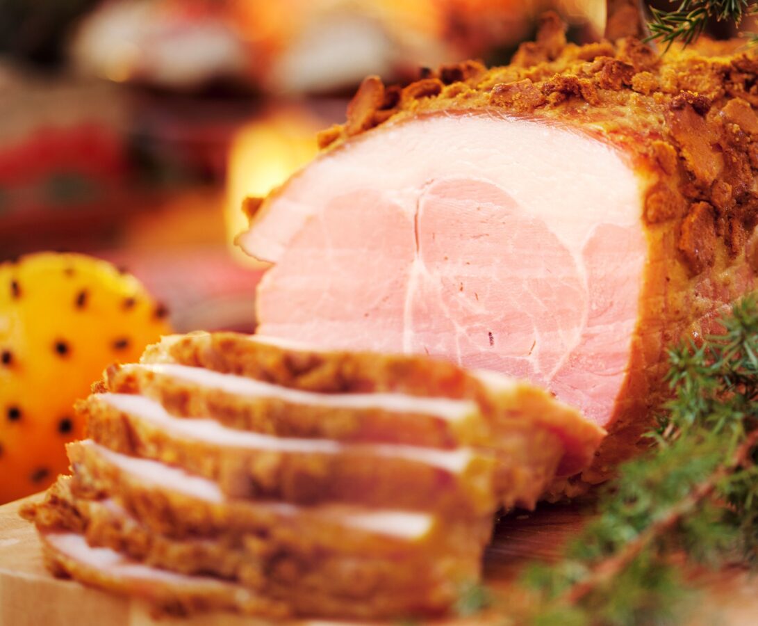 Skivad julskinka ligger upplagt mellan en nejlikedekorerad apelsin och grönkål