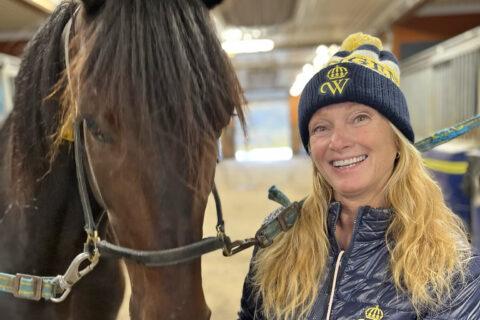 En kvinna i blå stickad mössa och jacka med Wångens emblem på står bredvid en mörkbrun häst i ett stall och båda tittar mot kameran.