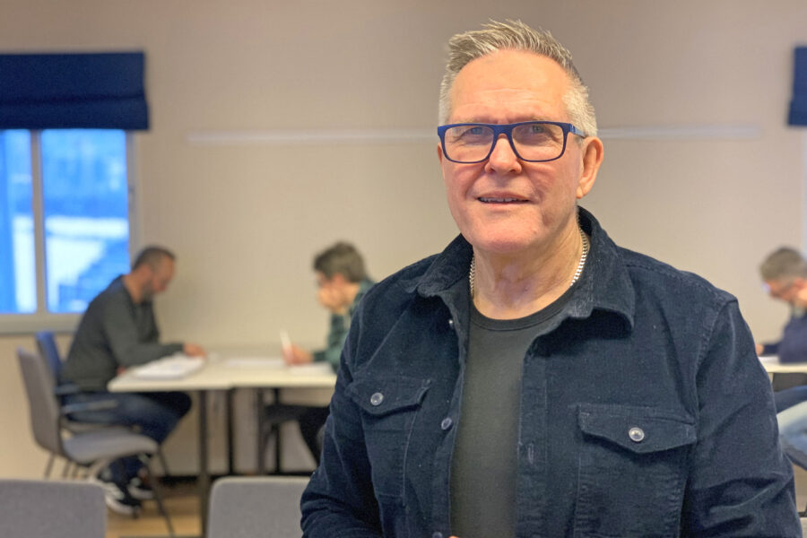 En man med glasögon och mörk kavaj håller i en kopp och ler mot kameran; i bakgrunden sitter människor och arbetar vid bord.