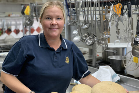 Madeleine Andersson, klädd i en mörkblå pikétröja, står i ett kök och håller i en bricka med stora, runda brödlimpor. I bakgrunden hänger diverse köksredskap.